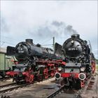 Links „011 100“, rechts „011 075-9“. Zwei schwarze Dampflokomotiven mit roten Fahrgestellen auf Bahngleisen.