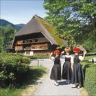 Drei Personen in traditioneller Tracht mit roten Hüten vor einem großen Fachwerkhaus mit blumengeschmücktem Balkon.