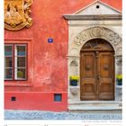 Das Hausnummernschild zeigt "77". Darunter ist ein rotes Gebäude mit verzierter Holztür und Pflanztöpfen.