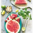 Text: Juli. Ein Tisch mit geschnittenen Wassermelonen, Limetten und Kräutern. Holzbrett mit Messer.