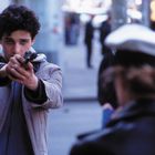 Ein junger Mann zielt mit einer Pistole auf eine Person in Polizeiuniform auf einer belebten Straße.