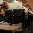 Ein Mann sitzt in einem Zugabteil und schaut aus dem Fenster, hält ein Handy, trägt einen Pullover; Haken mit Jacke.