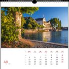 Kalenderblatt für Juli. Ein malerischer See mit einem großen Baum im Vordergrund und einem historischen Gebäude am Ufer.