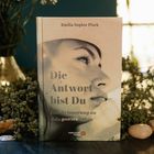 "Die Antwort bist Du. Eine Erinnerung an dein ganzes Leben." Buch von Emilia Sophie Plock. Gesicht einer nachdenklichen Frau.