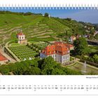 Juli, Weingut Schloss Wackerbarth. Ein Schloss mit roten Dächern, symmetrischer Garten und Weinberge im Hintergrund.