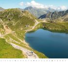 "Juli", Kalenderblatt mit Berglandschaft, Radfahrer auf einem Serpentinenweg, blauer Bergsee, im Hintergrund Berge.