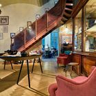 Ein Raum mit Holzboden, Wandbildern, Treppe, ausgestellten Schuhen auf Tisch, roten Sofas und einem Kronleuchter.