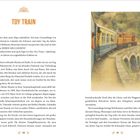 Titel: "Toy Train". Beschreibung des Zugs als Verbindung zwischen Welten und technisches Wunderwerk im Landschaftsbild. Bild zeigt einen Zug auf einem kurvigen Bahngleis über einer Brücke.