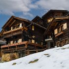 Holzhäuser mit Balkonen, Schnee bedeckt den Hang, blauer Himmel und einige Wolken im Hintergrund.