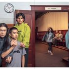 Oberhalb der Tür ist ein unscharfer Text. Personen in grauen und grünen Hoodies, eine läuft durch einen Flur.