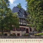 Kalendarblatt für Juli: Ein traditionelles Fachwerkhaus umgeben von grünen Bäumen und Blumenkästen.