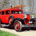 Nummernschild "1931", altes rotes Feuerwehrauto mit Holzleiter, große runde Scheinwerfer, blühende Bäume im Hintergrund.