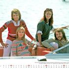Text: "Juli". Darunter ein Kalender. Vier Personen lächeln entspannt auf einem Boot am Wasser.