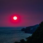 Ein leuchtend roter Sonnenuntergang über dem Meer, mit dunkler Küstenlinie und Himmel in tiefem Blau und Violett.