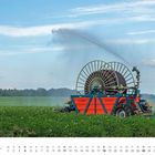 Kalenderseite Juli. Ein Bewässerungssystem in einem grünen Feld sprüht Wasser in die Luft.