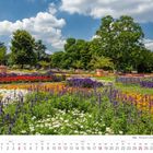 Kalendermonat Juli 2027. Farbenfrohe Blumenbeete im Park, umgeben von üppigen Bäumen und blauem Himmel mit Wolken.