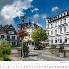 Text: "Wermelskirchen - Markt", "7", "2027". 
Eine malerische Stadtansicht mit traditionellen Gebäuden, Blumenampeln und blauem Himmel.