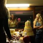 Keine erkennbaren Texte. Fröhliche Personen an einem Tisch mit einer großen Torte und Desserts. Eine Person hält einen Ball.