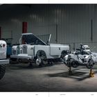 Text: "LAND", "CHAPTER 03", "BUILD".  

Ein Auto mit offener Motorhaube neben einer Karosserie auf Stützen.