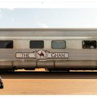 „Die wilde Kante“, „Australien“, „The Ghan“. Ein Zugwaggon mit einem Emblem. Ein Mann mit Hut geht daran vorbei.