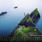 "Dingle Peninsula – wilde Atlantikküste." Steile, grasbewachsene Klippen erstrecken sich ins blaue Meer, umgeben von kleineren Felsen.