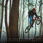 Kalender zeigt August, darunter Wochentage. Ein Mountainbiker springt in einem nebligen Wald über Felsen.