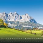Text: "Wilder Kaiser". Kalenderdaten unten. Grüne Hügel, Bäume, Alpenhütten, im Hintergrund dramatischer Berg.