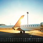 Kalender mit Datum und Wochentagen unten. Ein Flugzeug steht auf dem Rollfeld bei Sonnenaufgang.