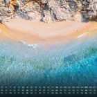 Kalenderdaten in weißem Text unten. Zu sehen ist ein Strand mit goldenem Sand, blauen Wellen und einer felsigen Küste.