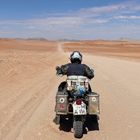 Nummernschild: "WM A 114". Ein Motorradfahrer fährt auf einer einsamen, staubigen Straße durch eine weite Wüstenlandschaft.