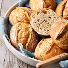 Einige runde, goldene Brötchen liegen in einer Schale auf einem blauen Tuch, daneben ein Messer auf Holz.