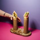 Zwei Wraps in Papprollen auf einem ovalen Tablett, umgeben von Nüssen. Eine Hand ist nahe an einem Wrap. Hintergrund rosa/lila.