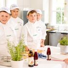 Fünf Kinder in weißen Kochoutfits mit "MINIKÖCHE"-Mützen arbeiten an einer Küchentheke mit Gewürzen und Zutaten.