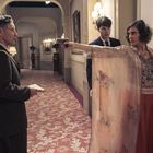 Eine Frau zielt mit einer Pistole auf einen Mann in einem eleganten Korridor, eine weitere Person steht dahinter.