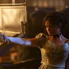 Eine Frau in einem eleganten Kleid zielt mit einer Pistole. Im Hintergrund sind Pferdekutschen zu sehen.