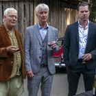 Drei Männer in Anzügen halten Gläser Wein. Sie stehen im Freien vor einer Holzhütte und einem Tisch mit Weinflaschen.