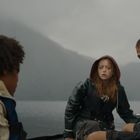 Drei Personen in Regenkleidung sitzen in einem Boot auf einem nebligen See, umgeben von Bergen.