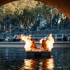 Zwei Figuren in einem brennenden Boot unter einer Brücke in einer Stadt mit Bäumen und Menschen auf einem Boot im Hintergrund.