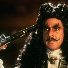 Ein Mann mit lockigem Haar hält eine Pistole an den Kopf. Er trägt einen Schnurrbart und elegante Kleidung mit Brosche.