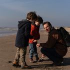 Drei Menschen am Strand; ein Erwachsener mit Rucksack hockt, zeigt zwei Kindern in Jacken etwas im Sand. Am Horizont Meer.