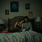 An der Wand ein Poster mit "PLAYERS". Zwei Personen sitzen auf einem Bett, sie lehnt sich an ihn. Im Raum stehen Lampen.