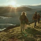 Menschen wandern bei Sonnenuntergang über eine moosige Landschaft, neben einem Gletscherfluss.