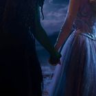 Zwei Personen halten Hände; eine trägt ein schimmerndes, helles Kleid, die andere hat dunklere Kleidung und grüne Haut.