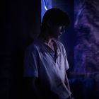 Ein Junge in einem schmutzigen, weißen Hemd lehnt in einer düsteren Umgebung an einer Wand, in blauem Licht getaucht.