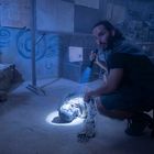 Eine Person hält eine Taschenlampe und leuchtet auf eine am Boden liegende, mumifizierte Gestalt in einem düsteren Raum.