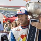 Text "WINNER" auf einer Trophäe. Ein Mann in Rennanzug mit "Wonder" Logo hält die Trophäe, umgeben von Menschen.