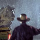 Ein Dinosaurier im Regen steht einem Mann mit Hut gegenüber, der eine rote Fackel in der Hand hält.