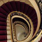 Eine Wendeltreppe mit rotem Teppich und kunstvollen Geländern von oben betrachtet, mit einer Person, die hinaufgeht.