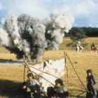 Menschen laufen auf einer Wiese vor einer Explosion davon, während zwei Reiter im Hintergrund galoppieren.