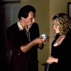 Ein Mann hält Geldscheine einer Frau entgegen. Der Raum ist warm beleuchtet, im Hintergrund ein Gemälde und eine Skulptur.